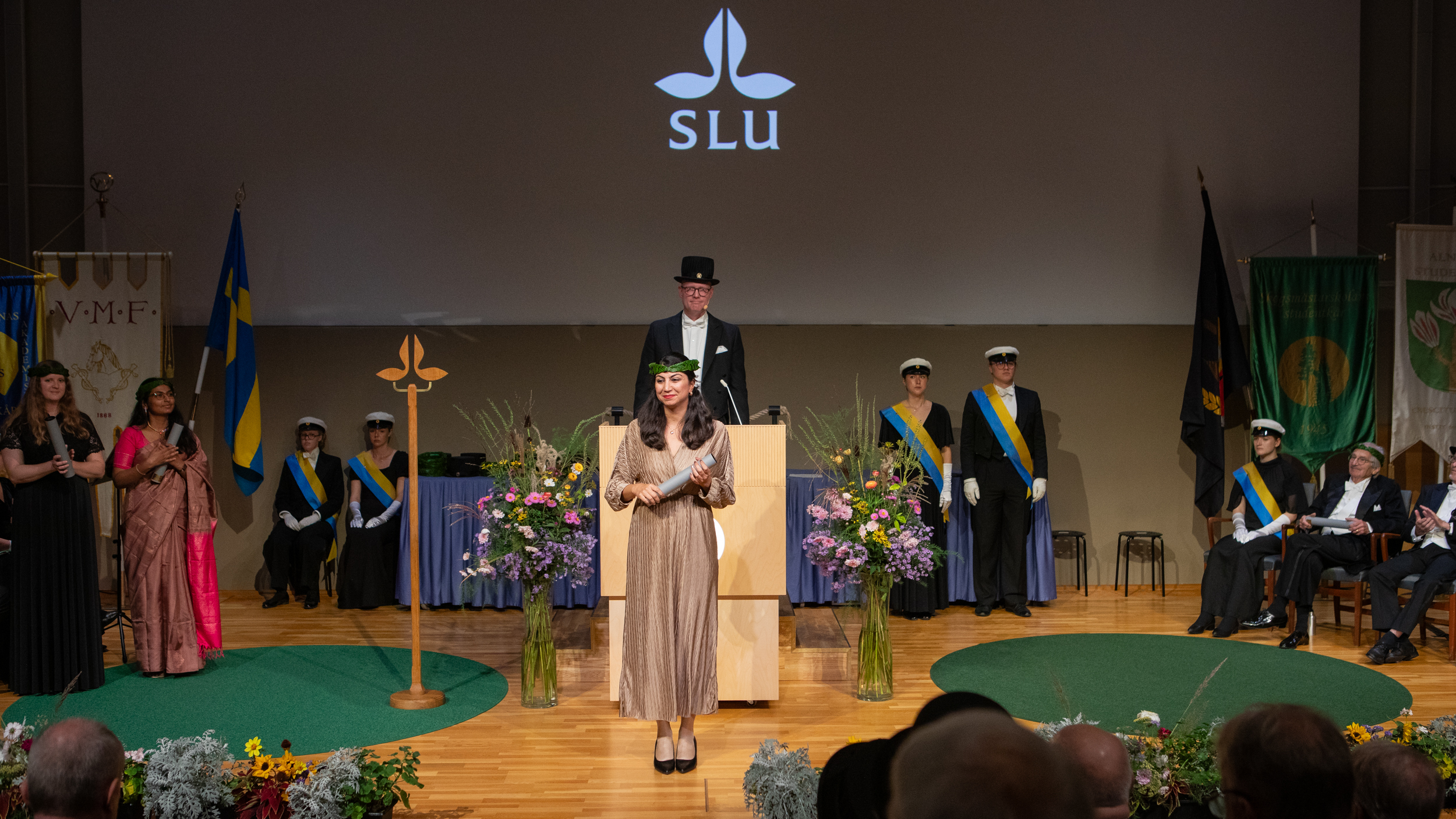 A person with long brown hair, wearing a brown dress and a laurel wreath, is standing on a stage holding a rolled paper.