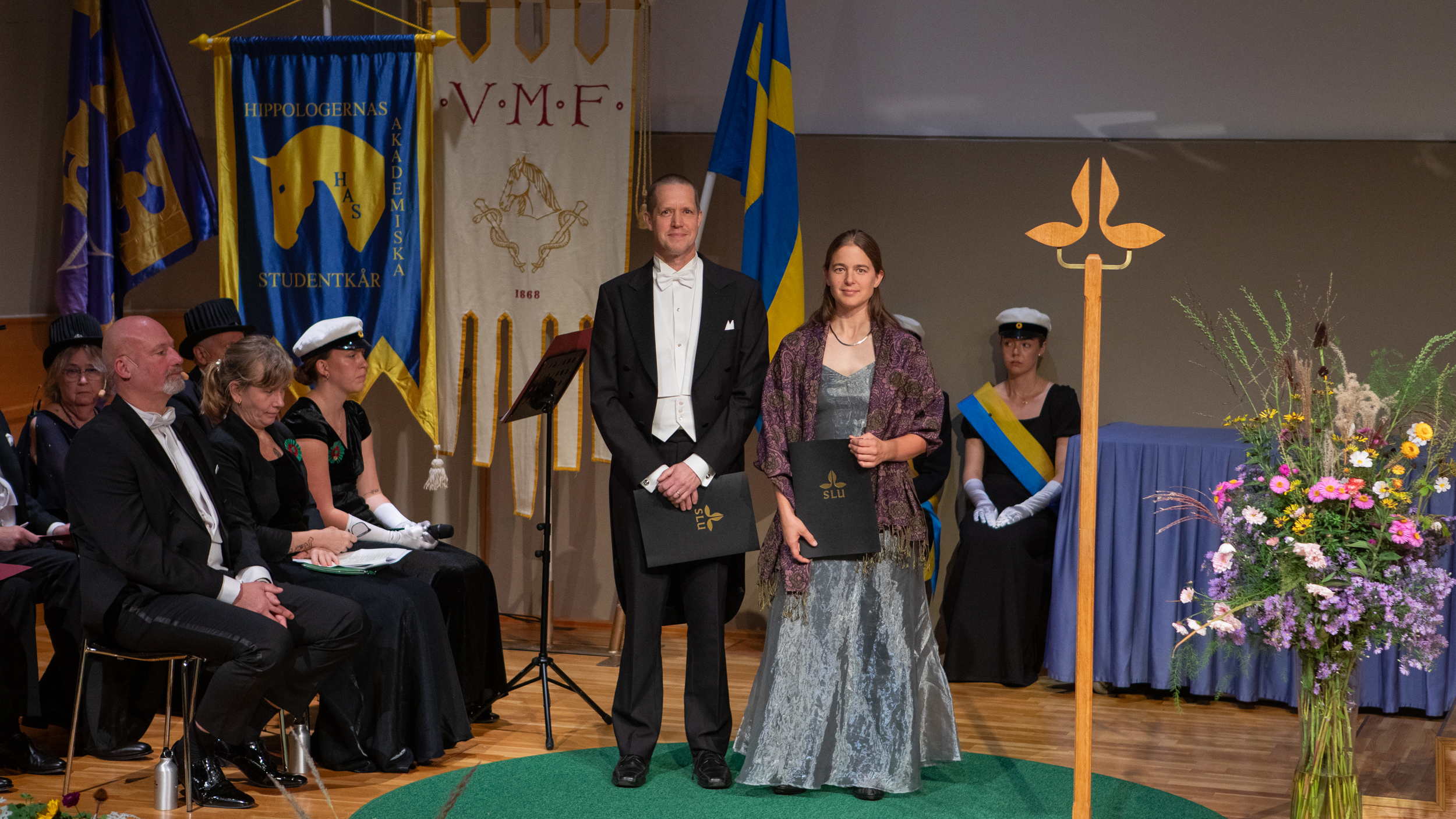Two people wearing white tie, standing on a stage holding black diploma folders.