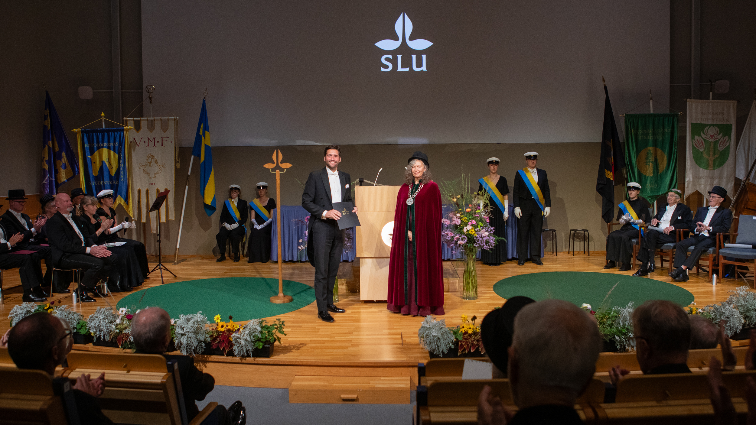 Two formally dressed individuals on a stage. One is holding a black folder bearing a logo, while the other is wearing a doctoral hat, a Vice-Chancellor’s chain and a red velvet gown.