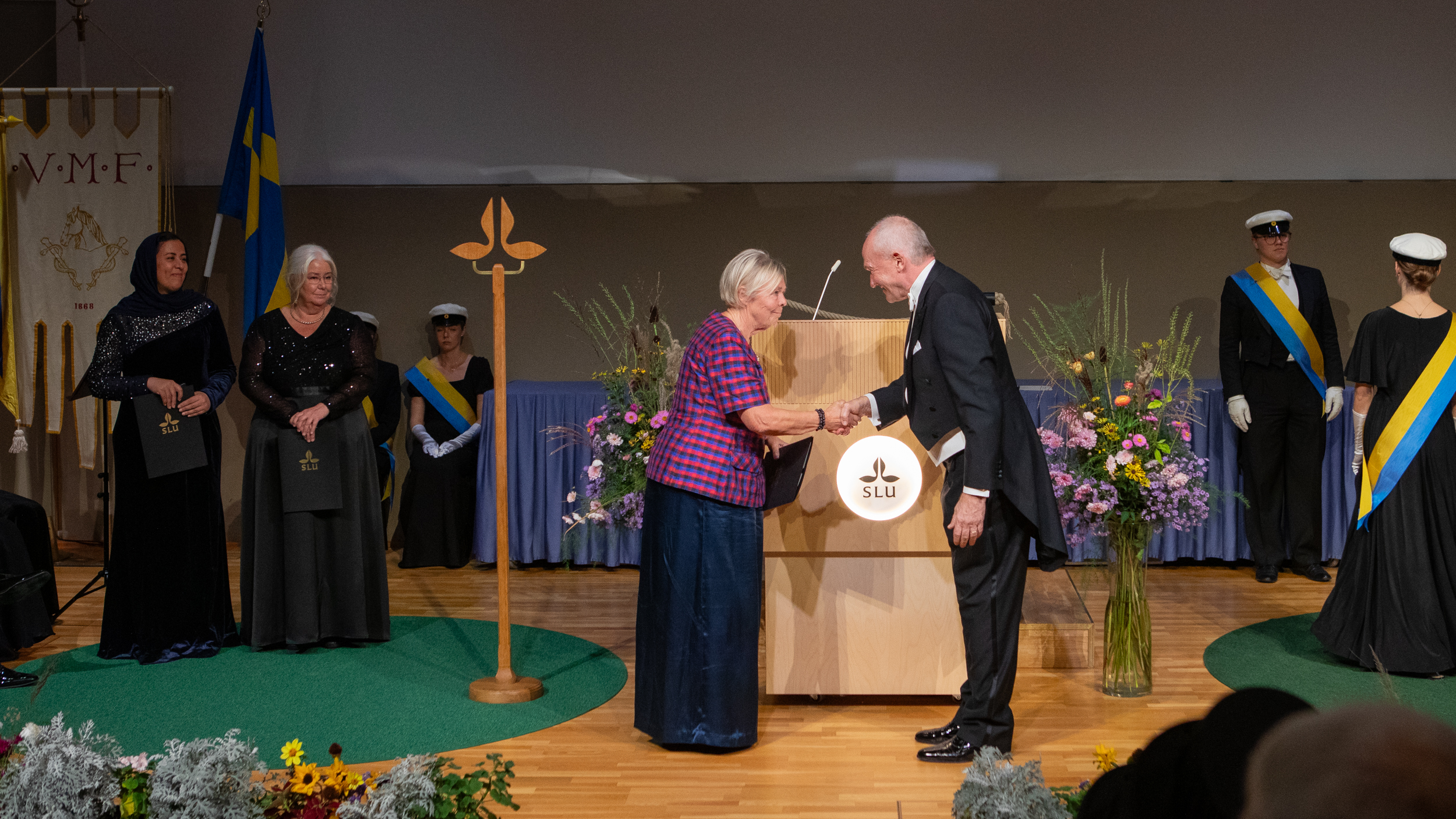 Two festively dressed individuals shaking hands on a stage. In the background there are two more individuals holding black diploma covers.
