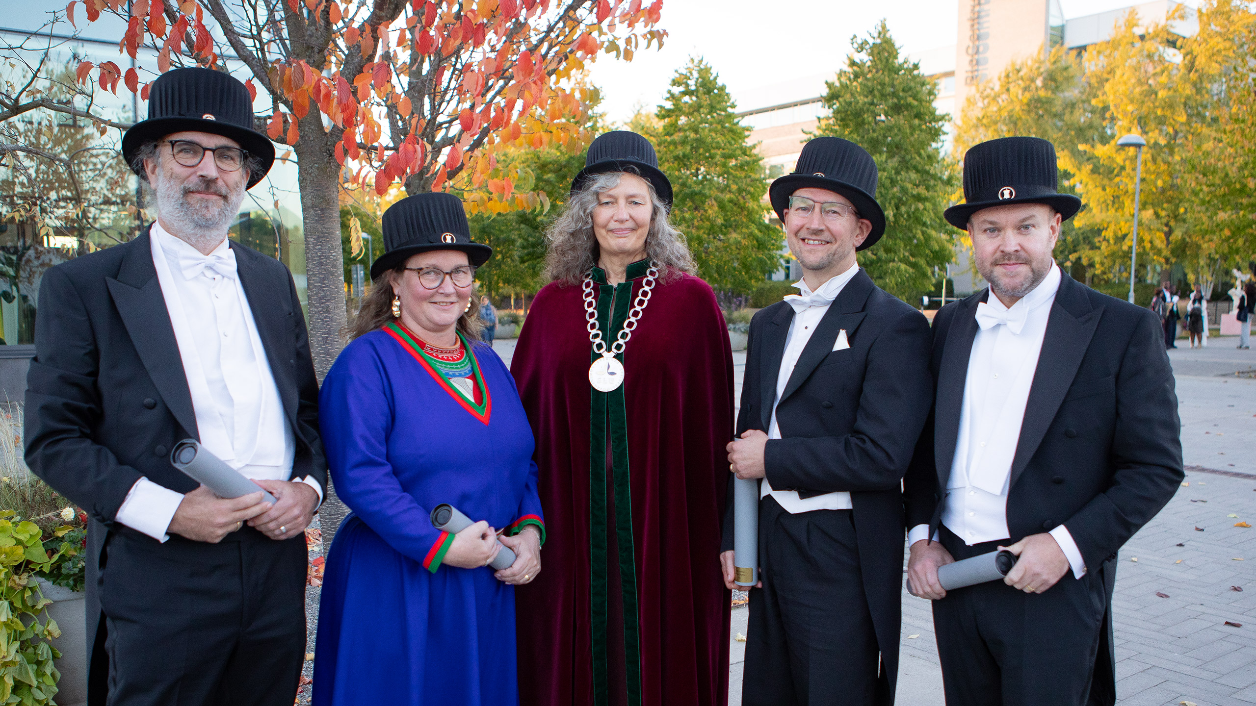 Five festively dressed persons wearing black doctoral hats standing outside.