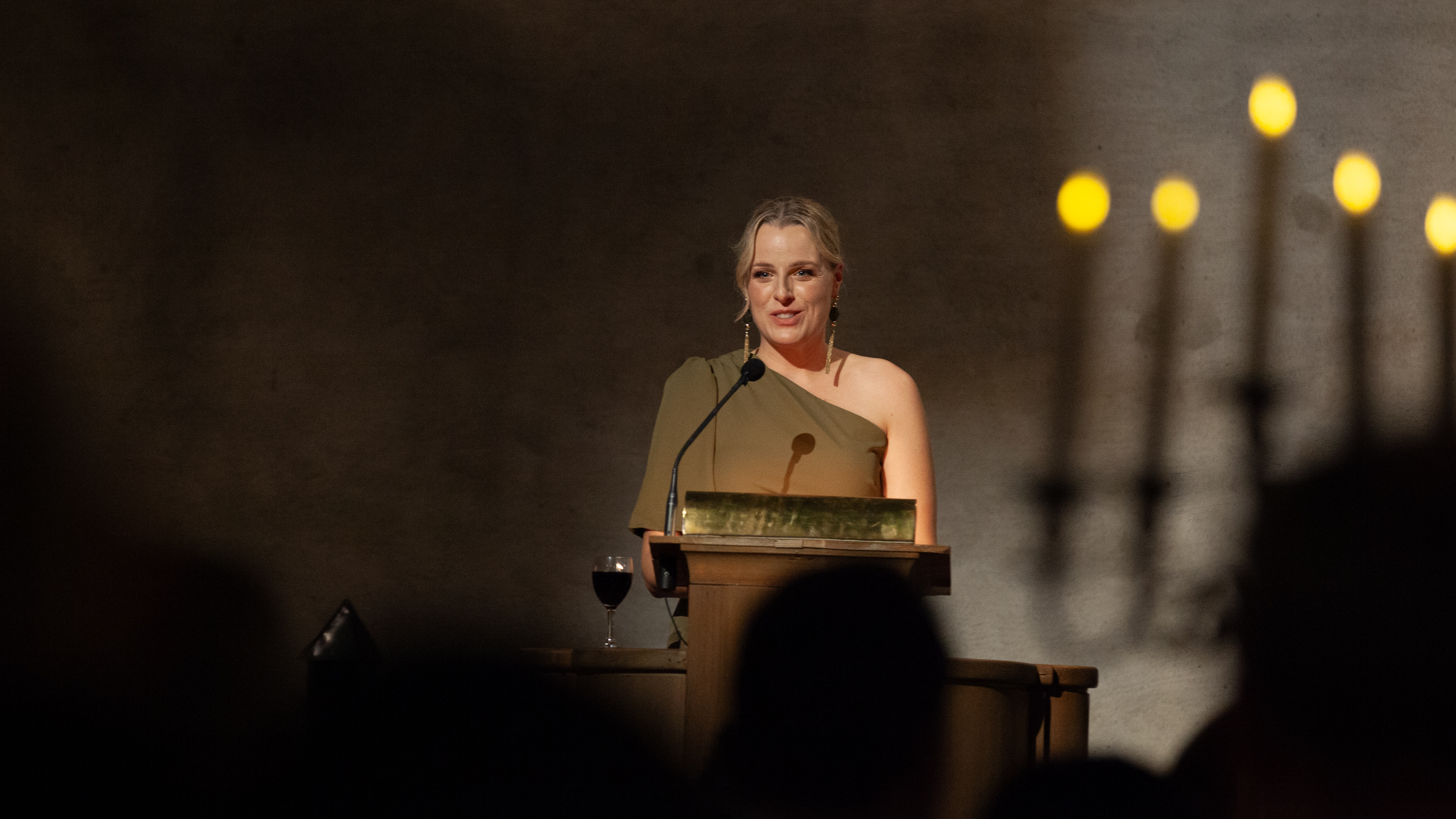 A person in a beige dress standing at a lectern smiling.