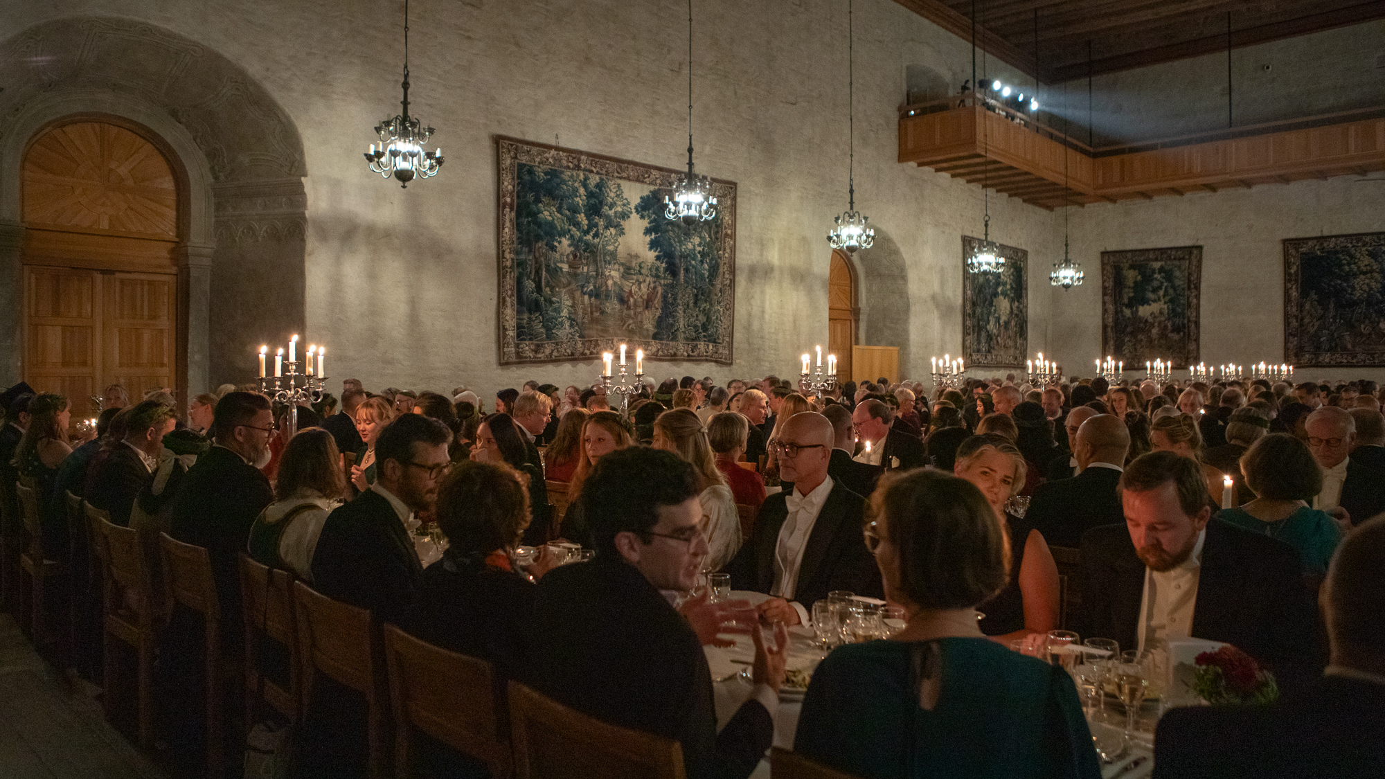 En stor gammal sal med middagsgäster vid en mängd långbord.