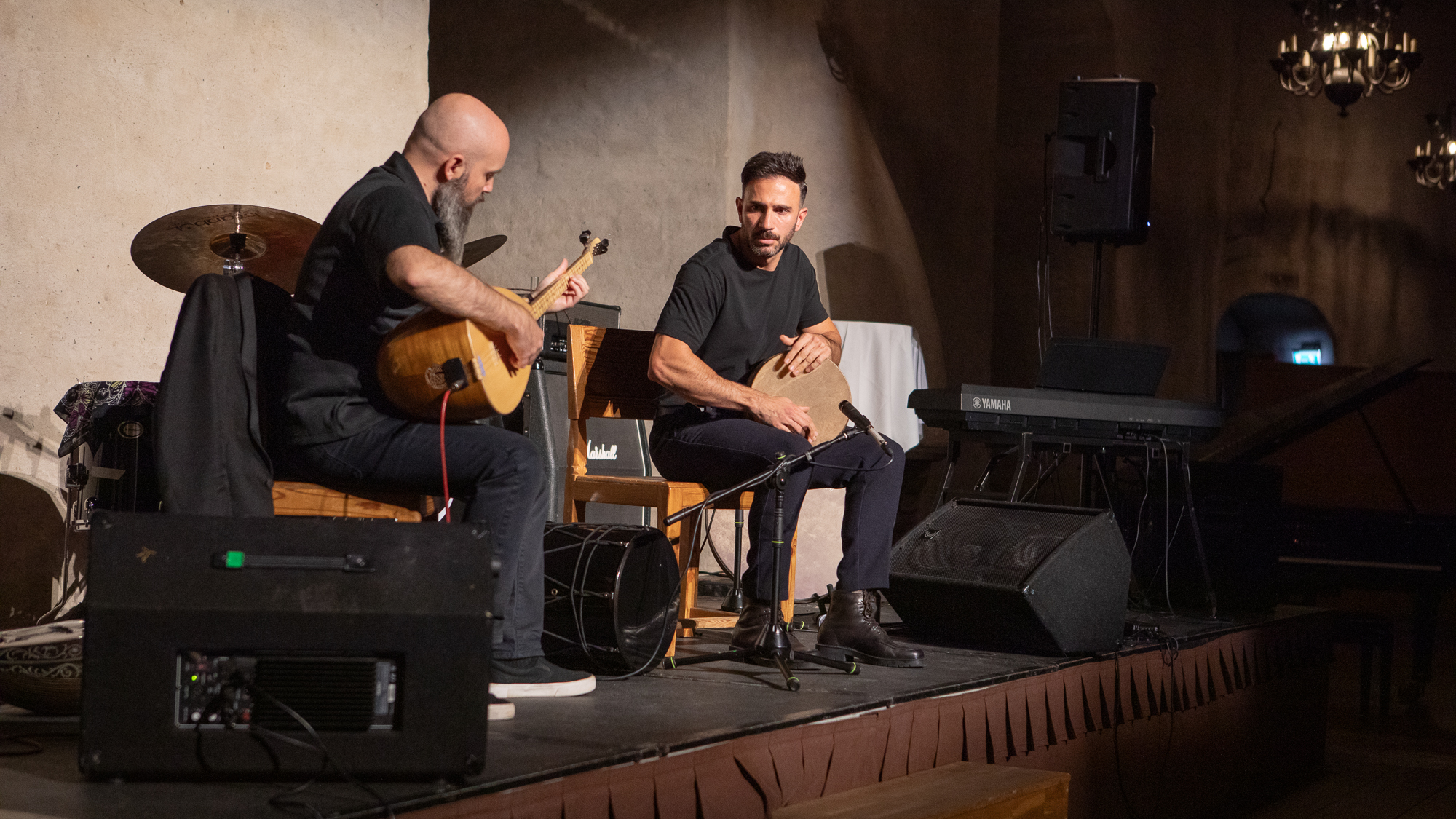 Två personer sitter på en scen, den ena hållandes en trumma, den andra något slags stränginstrument med rund kropp.