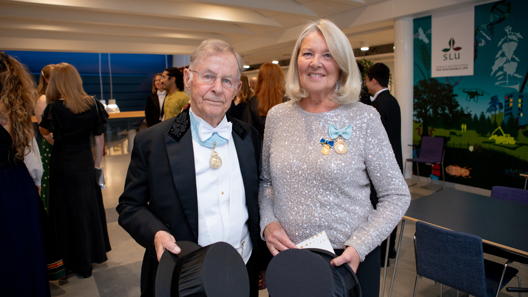 Två äldre personer i högtidsdräkt och med medaljer på bröstet står och håller i varsin doktorshatt. 