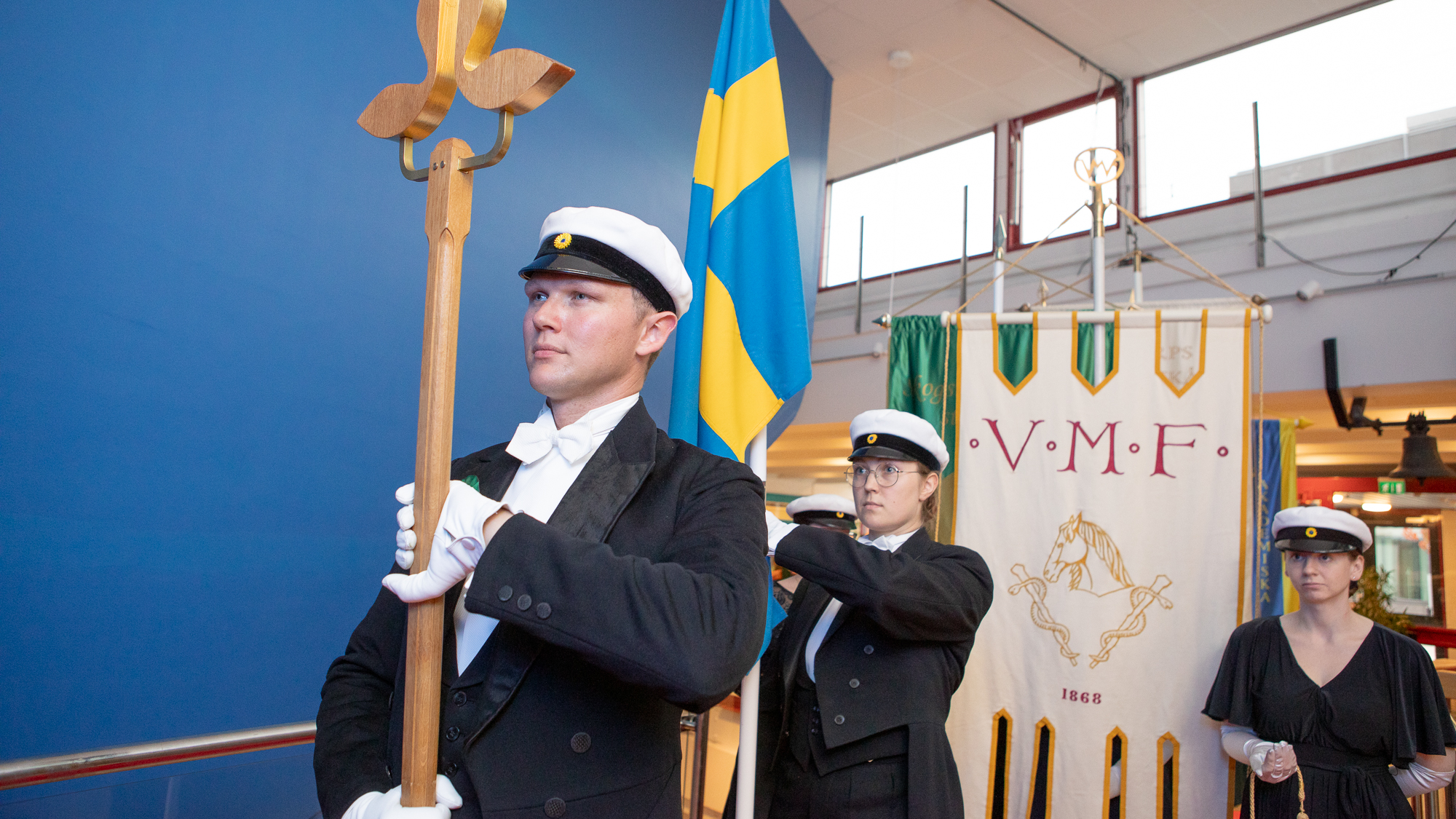 Studenter i frack och studentmössa står på rad. Den längst fram håller en trästav med en SLU-logotyp i toppen. De bakom håller i fanor. 