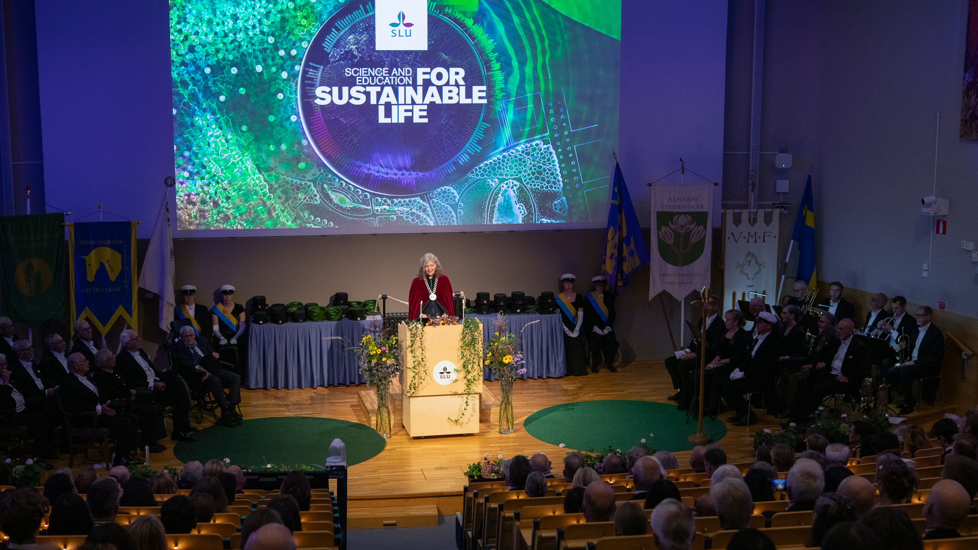 En kvinna iklädd röd sammetskåpa och stor halskedja i silver står i en talarstol på en scen, inför en publik. 