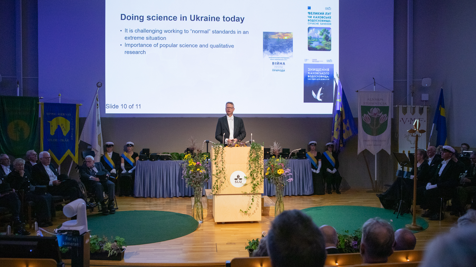 En man står i talarstolen på en scen. Bakom honom visas en projicerad presentation på väggen.