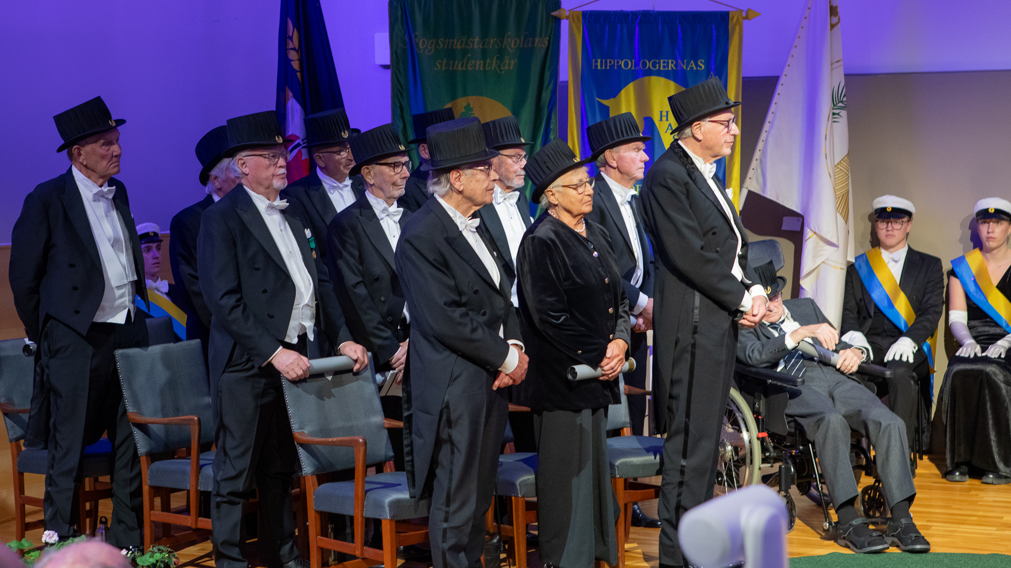 Tolv äldre högtidsklädda personer i doktorshatt står framför sina stolar på en scen.
