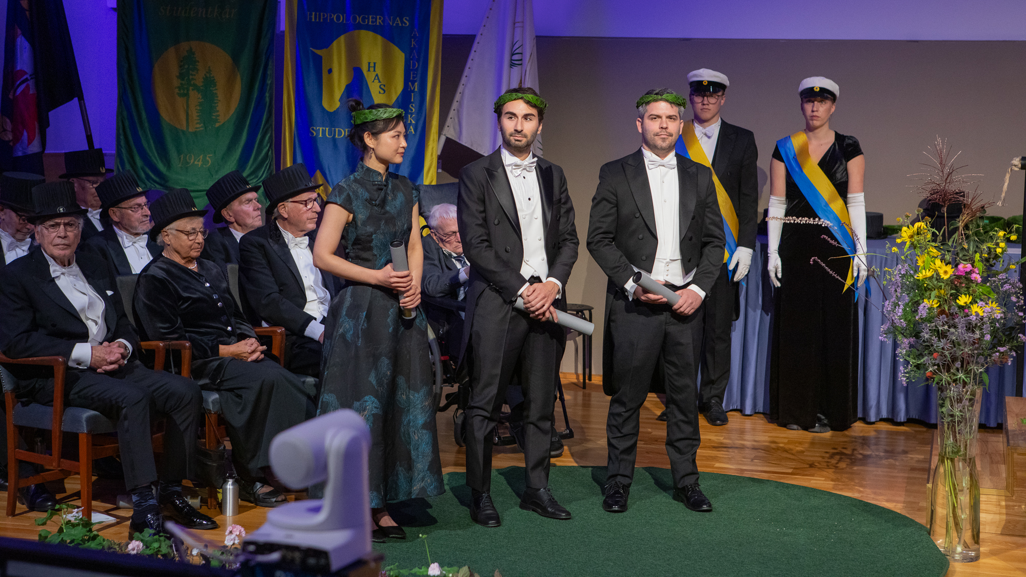 Tre personer i högtidskläder, med lagerkransar på huvudet, står på en scen. I bakgrunden sittande personer. 