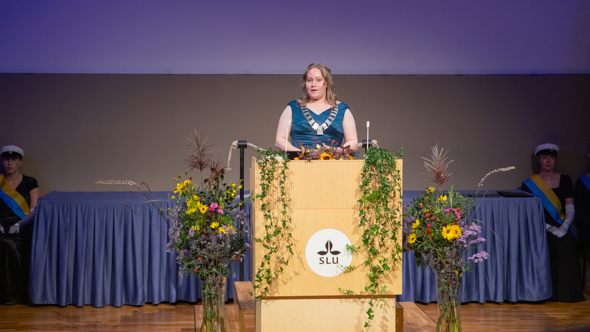 En ung person i stor halskedja och blå klänning står i en talarstol på en scen.