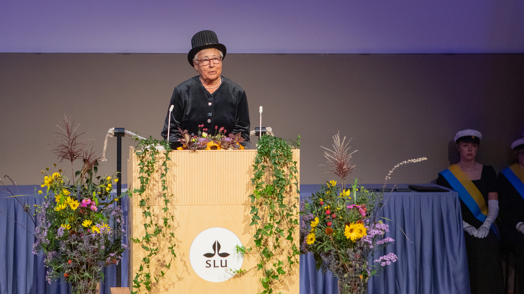 En äldre svartklädd kvinna i doktorshatt står i en talarstol. 