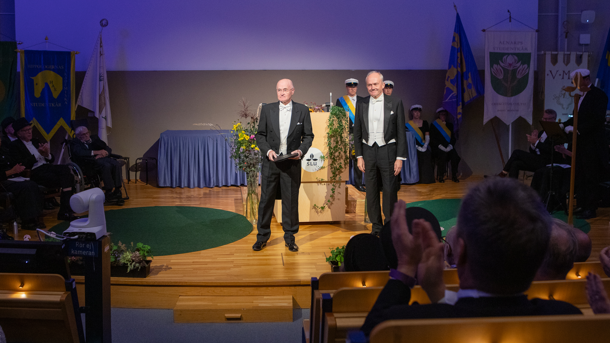 Två personer i frack står längst fram på en dekorerad scen. En av dem håller i en svart mapp med guldlogotyp.