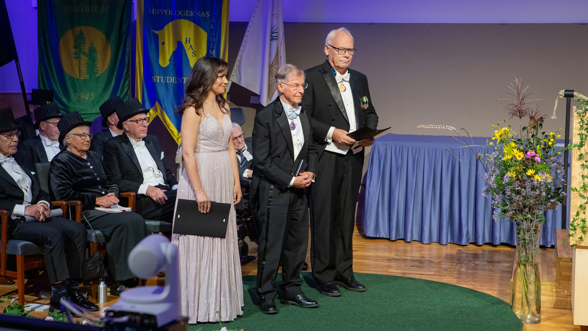 Tre personer står på en scen, en i klänning och två i frack, med diplom i handen.