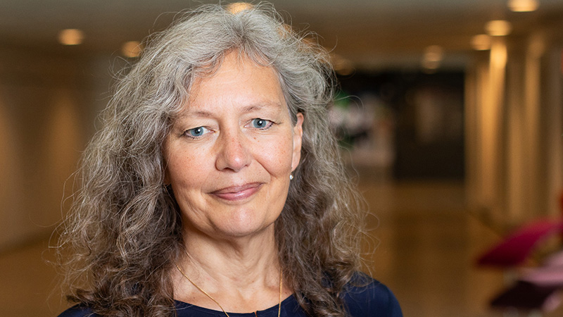 Leende kvinna med grått, lockigt hår står i korridor. Foto