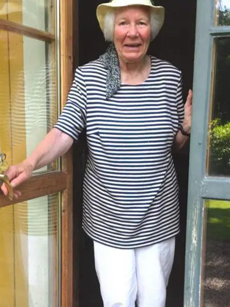 En äldre kvinna i hatt står i en dörröppning och ler.