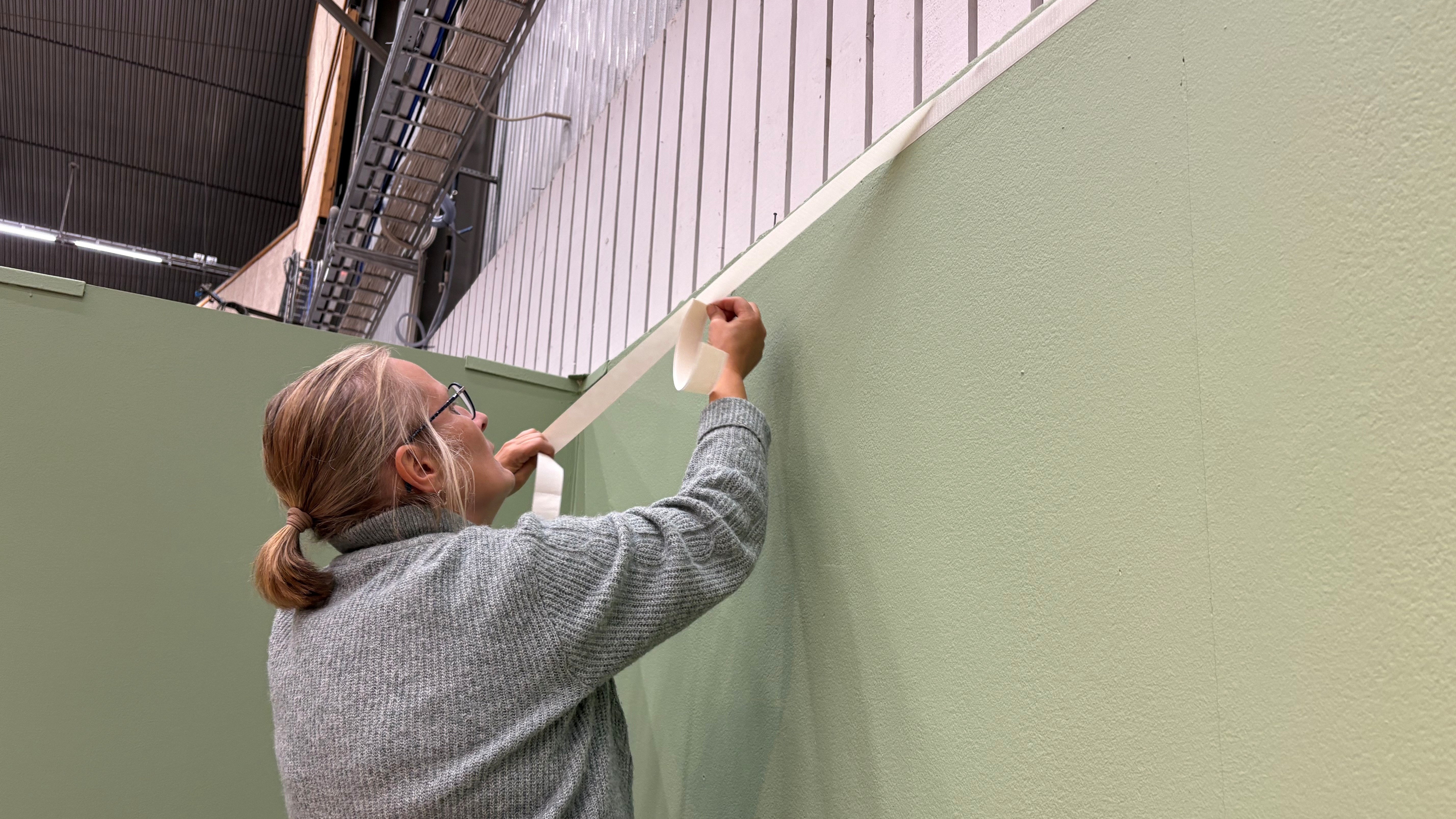 En kvinna klädd i grå tröja sträcker sig mot kanten av en montervägg. I ena handen håller hon en tejprulle och hon klistrar upp en tejpremsa överst på väggen. Väggen är ljust grön. 