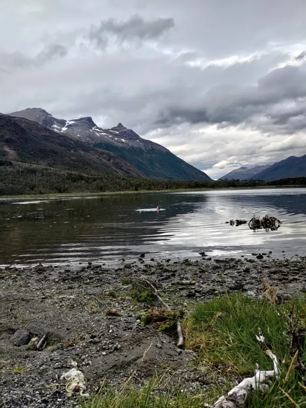 Person swimming in a calm lake surrounded by mountains under a cloudy sky.
