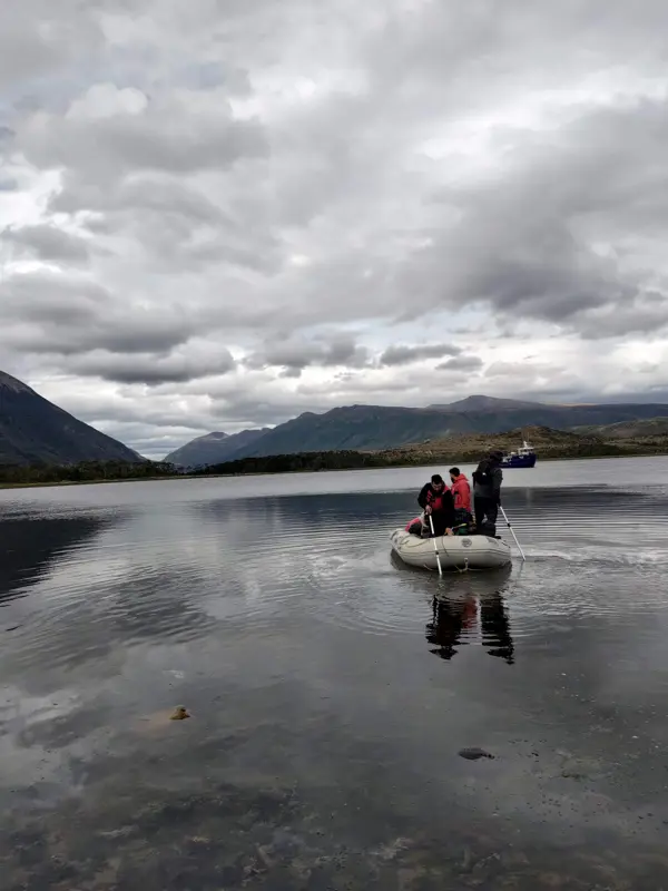 Small inflatable boat with people on calm water, with mountains and a ship in the background.
