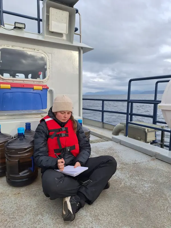 Person wearing a life jacket sits on a boat, writing in a notebook at sea.