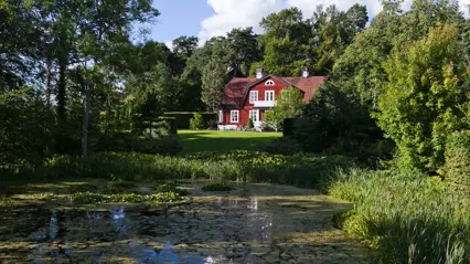 A red building surrounded by greenery with a pond in the foreground. Photo.