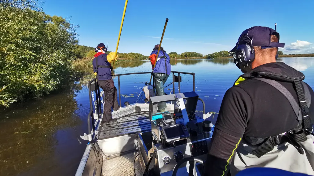 Three people in life jackets and hearing protection are boat fishing from an aluminum boat in a calm water area surrounded by greenery and blue sky. Two people stand in the bow with yellow rods in the water while a third steers the boat.