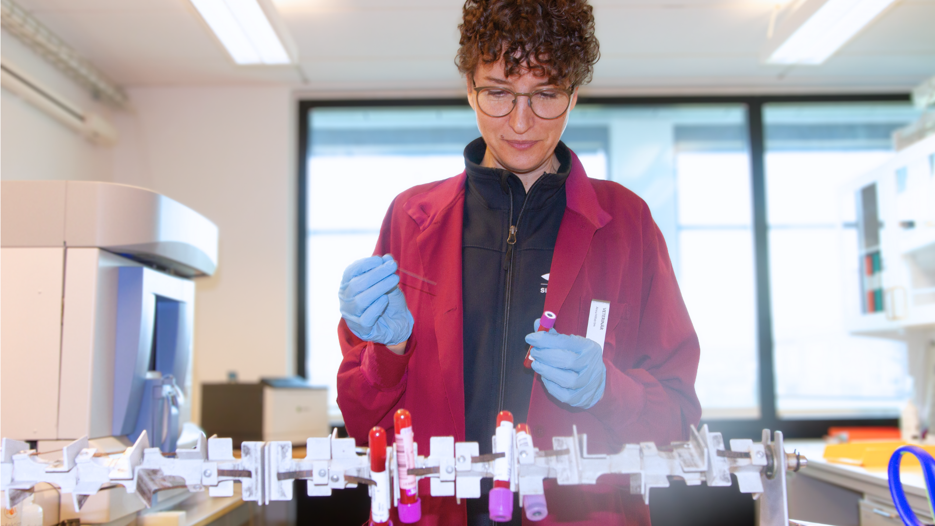 Person with short, brown curly hair and glasses, wearing burgundy clothes. In front of the person is a rack of test tubes with tubes of liquid inside.