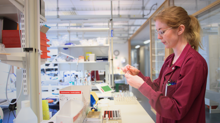 Kvinna med blont hår, glasögon och iklädd en röd rock som står upp. Framför henne är ett bord med labbutrustning. I bakgrunden syns hyllor med föremål. 
