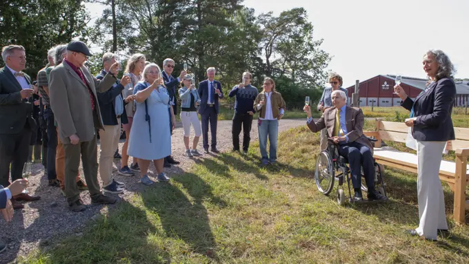 En äldre man i rullstol och en kvinna med champagneglas i händerna står framför en kohage och skålar med en mindre folksamling.