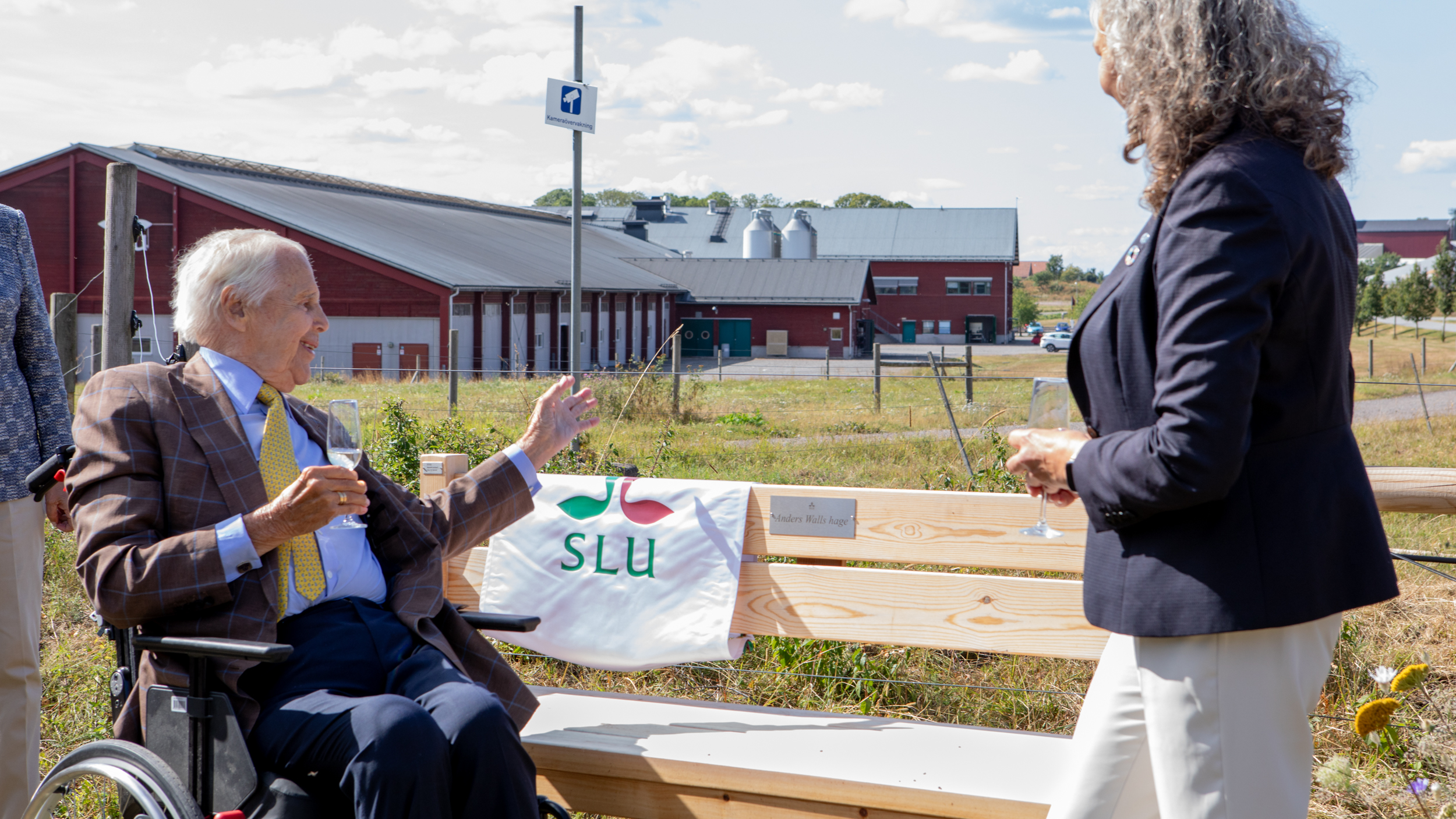 En äldre man i rullstol och en kvinna står framför en träbänk. Mannen drar en liten glansig duk med en logotyp åt sidan och blottar en plakett med texten "Anders Walls hage". I bakgrunden skymtar en kohage. 