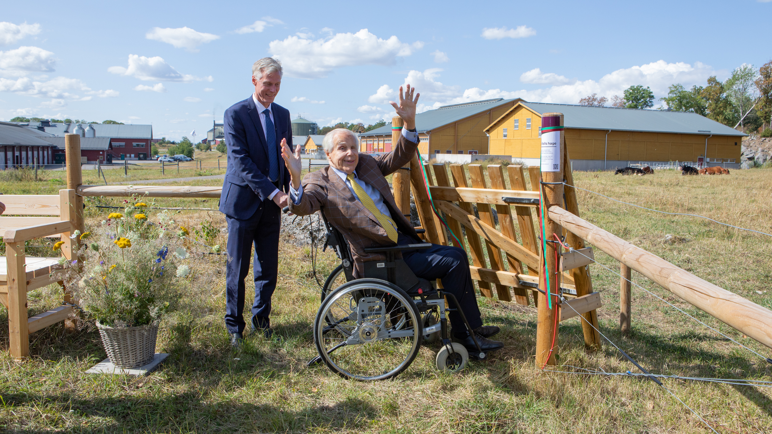 En man står bakom en rullstol vari en äldre man sitter. De befinner sig vid grinden till en kohage. Den äldre mannen ser glad ut och sträcker upp armarna.
