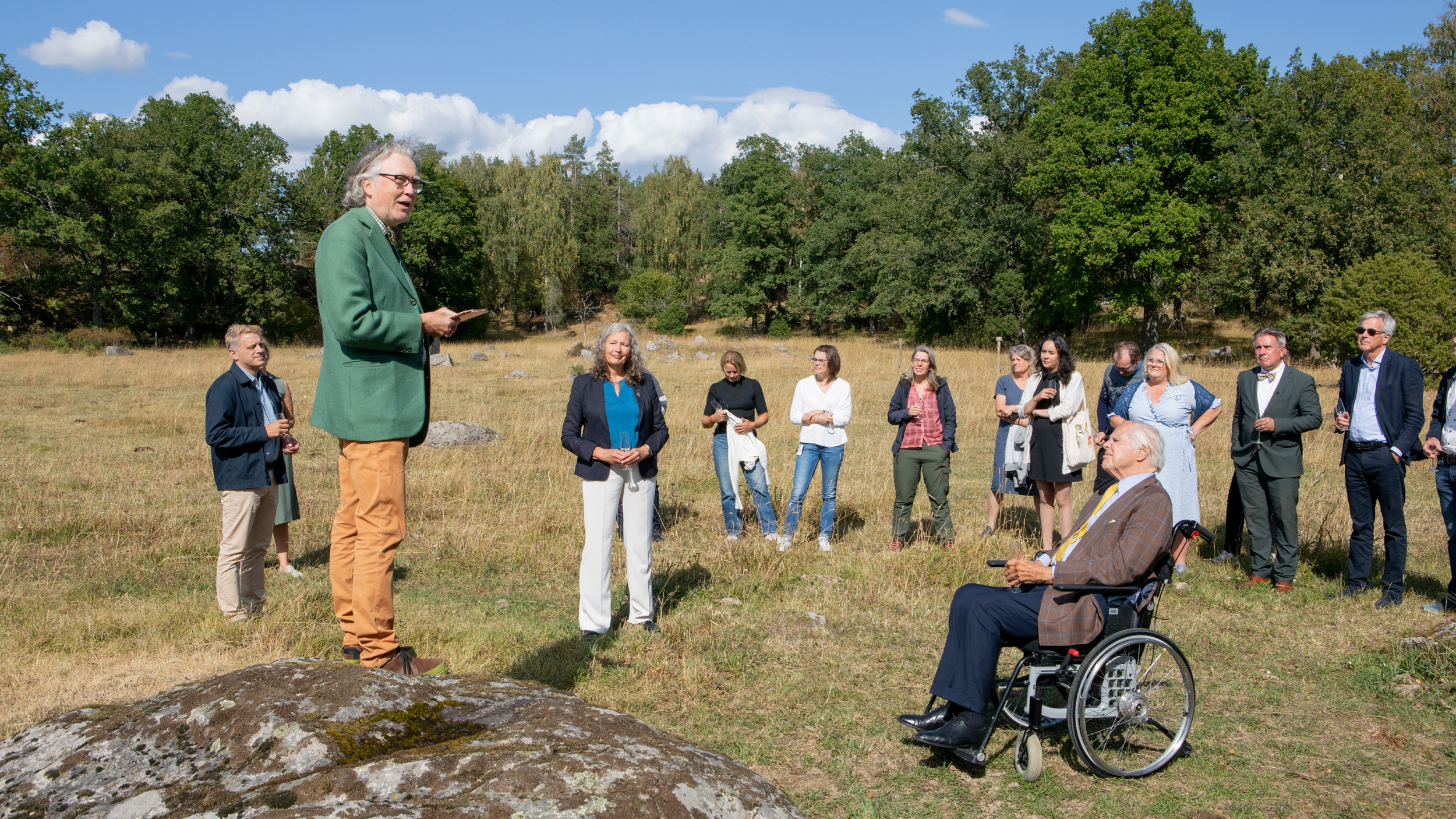 En person med grått långt hår, klädd i ljusbruna byxor och grön kavaj, står på en sten i en naturbeteshage och talar inför en publik.