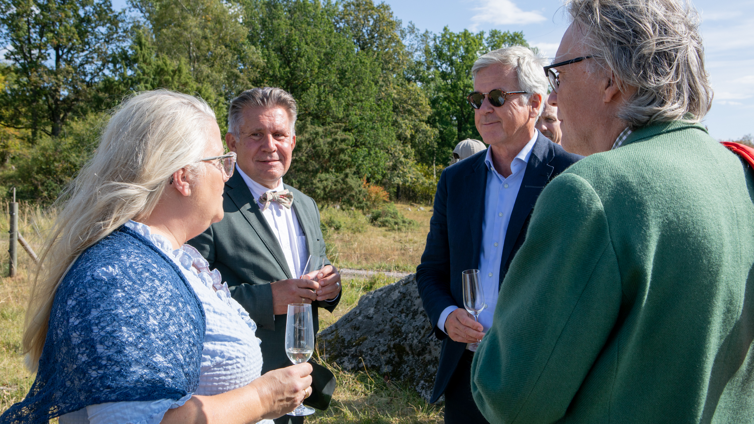 En konversation mellan fyra personer i en hage. En av dem klädd i ljusblå klänning, de resterande klädda i kavaj.