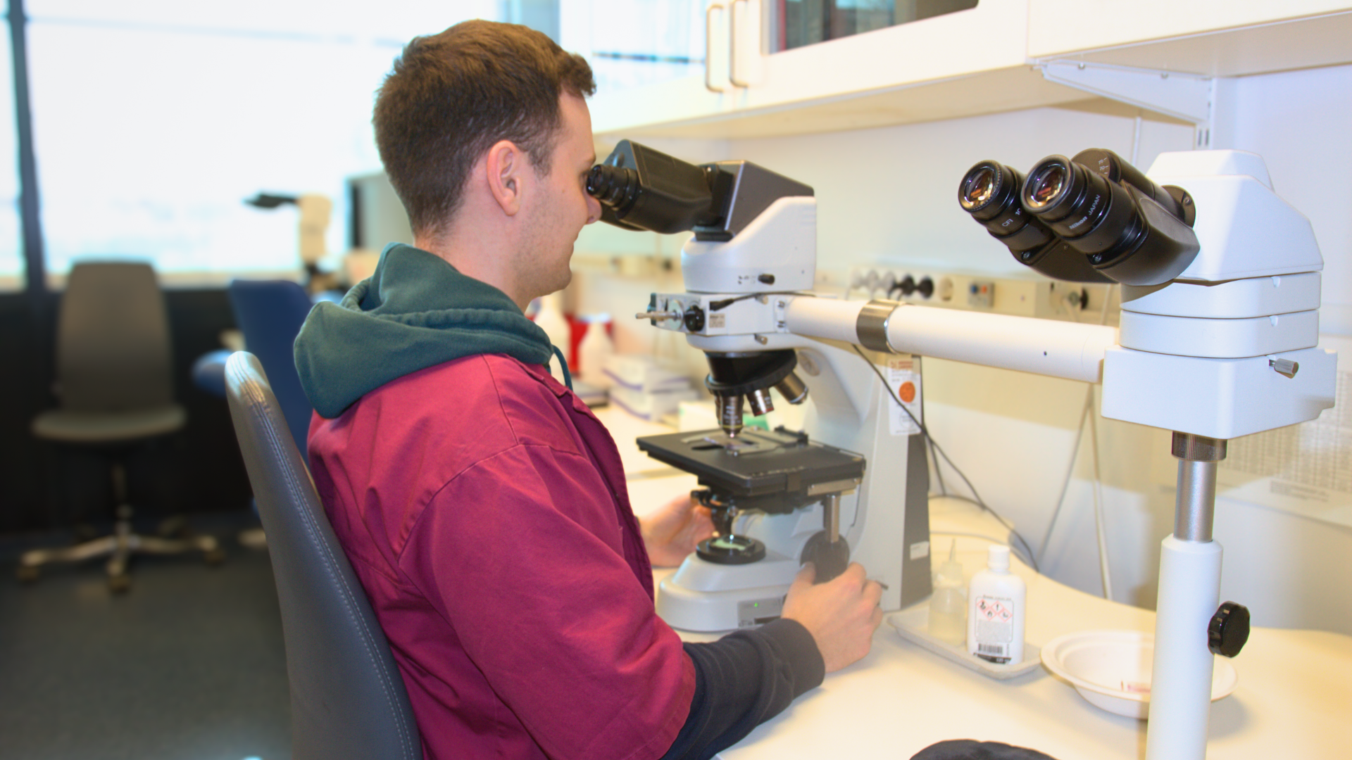 Man som sitter framför ett mikroskop. Mannen har kort brunt hår och har på sig en röd rock med mörkgrön huva.