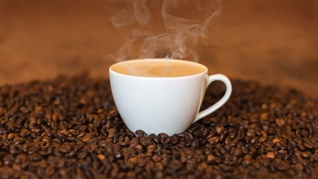 En rykande kopp kaffe omgiven av bruna kakaobönor. Foto: Pixabay
