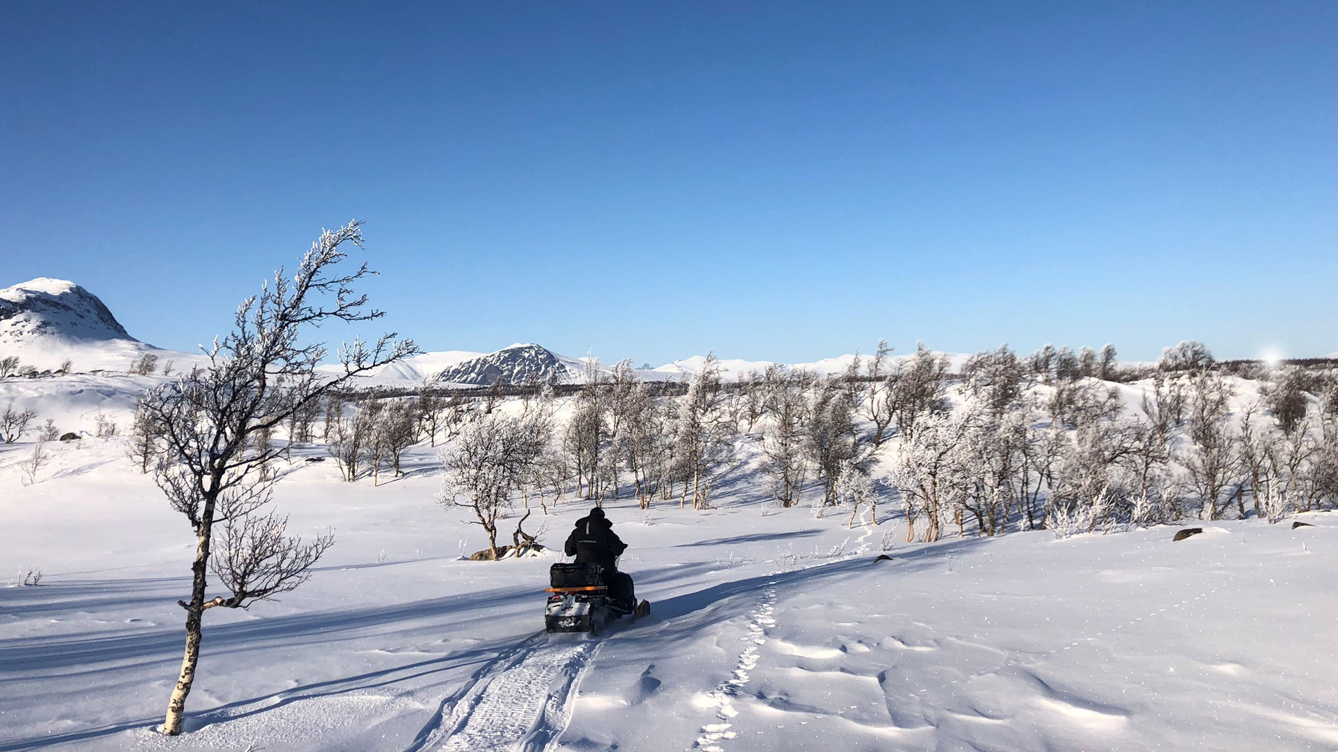 Snöskoter kör genom snötäckt fjällandskap under klarblå himmel.