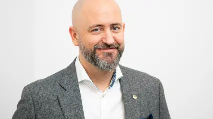 En person med kort, grått skägg och flint står mot en vit bakgrund. Personen bär en grå kostym med vit skjorta och en liten pin på kavajen.