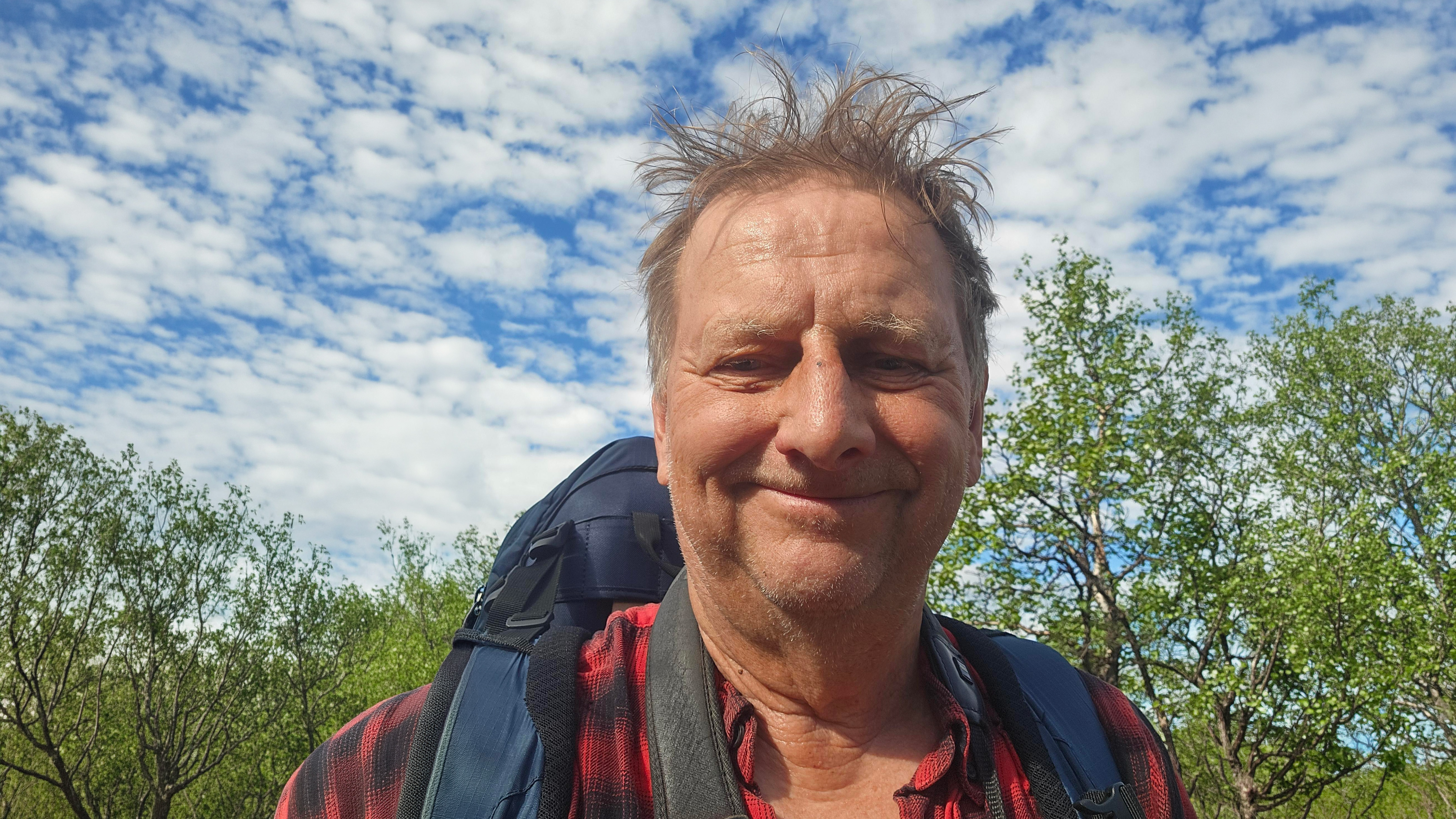 Porträttfoto av en person utomhus. Personen bär en mörk ryggsäck och en röd- och mörkrutig skjorta. I bakgrunden syns grönskande träd och en himmel med vita moln mot blå bakgrund.