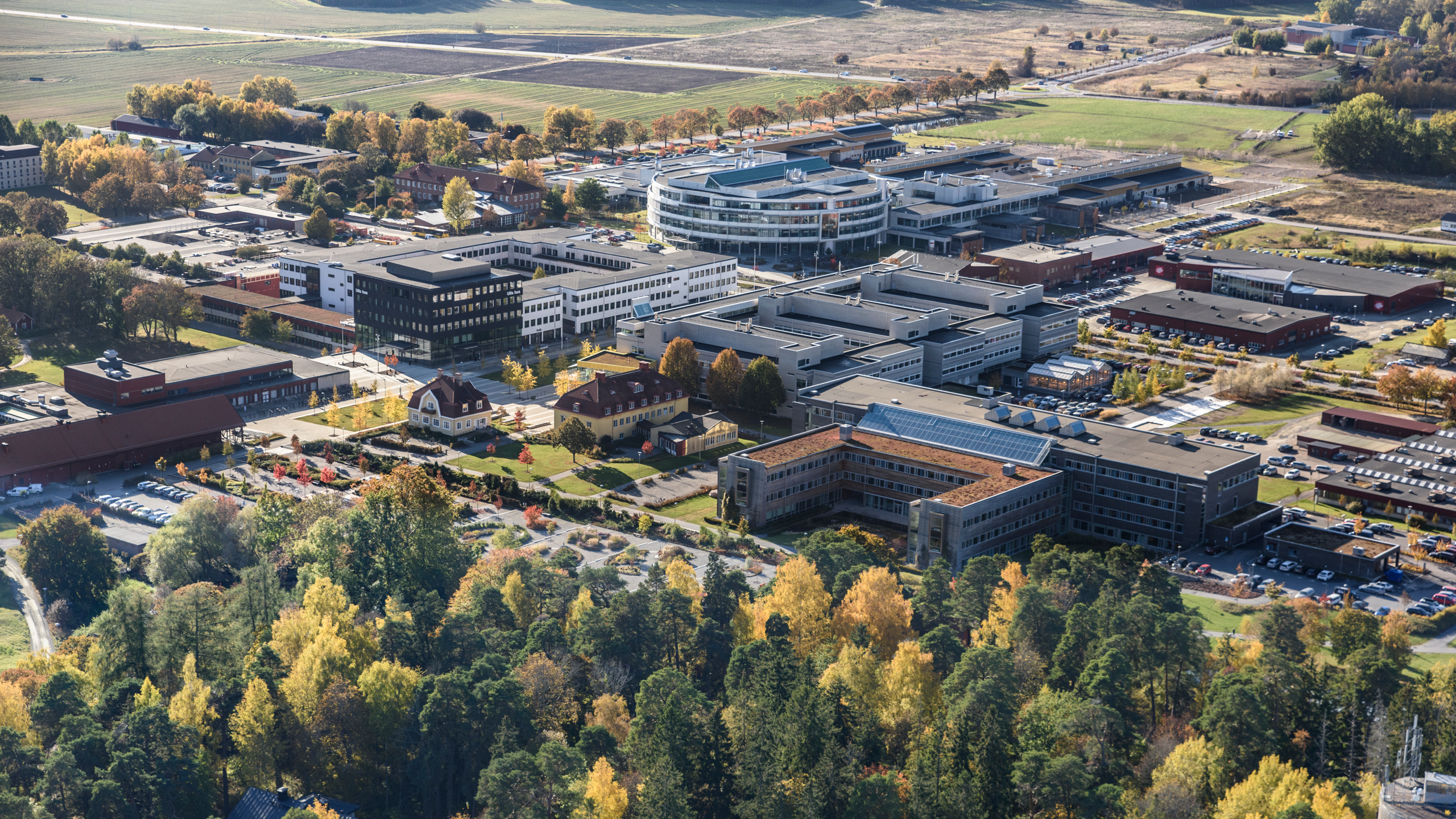 Flygbild över ett stort campusområde med flera moderna byggnader omgivna av höstfärgade träd. I centrum syns en rund byggnad med glasfasad, omgiven av rektangulära kontors- och institutionsbyggnader. Parkeringsplatser och vägar löper mellan byggnaderna, och i bakgrunden syns öppna fält och skog. Foto.
