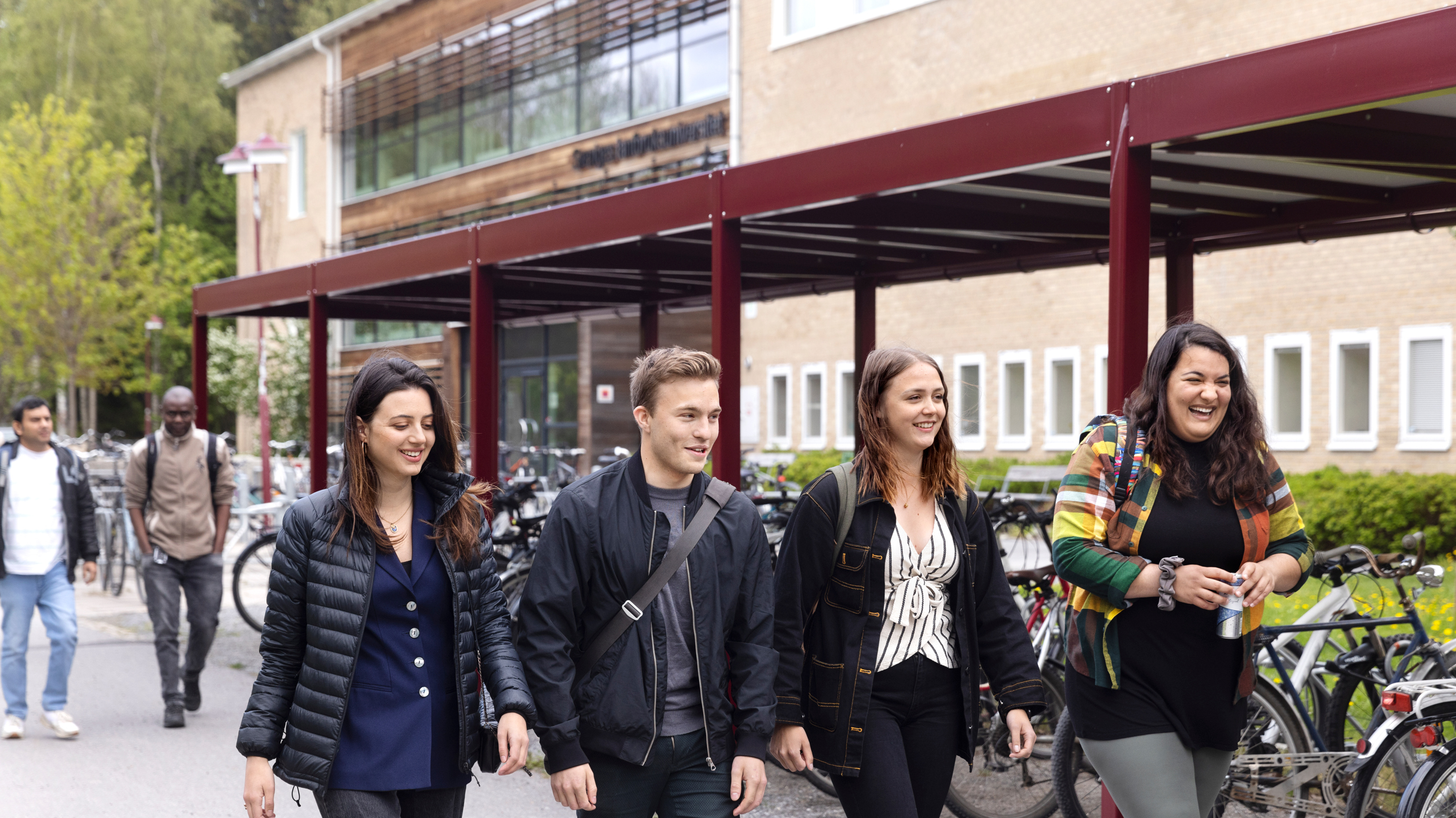 Grupp av personer som går längs en gångväg vid ett campusområde. I bakgrunden syns en byggnad med stora fönster och en röd metallkonstruktion som fungerar som cykelskydd. Flera cyklar står parkerade längs sidan, och gröna träd omger området. Foto: Malin Grönborg.