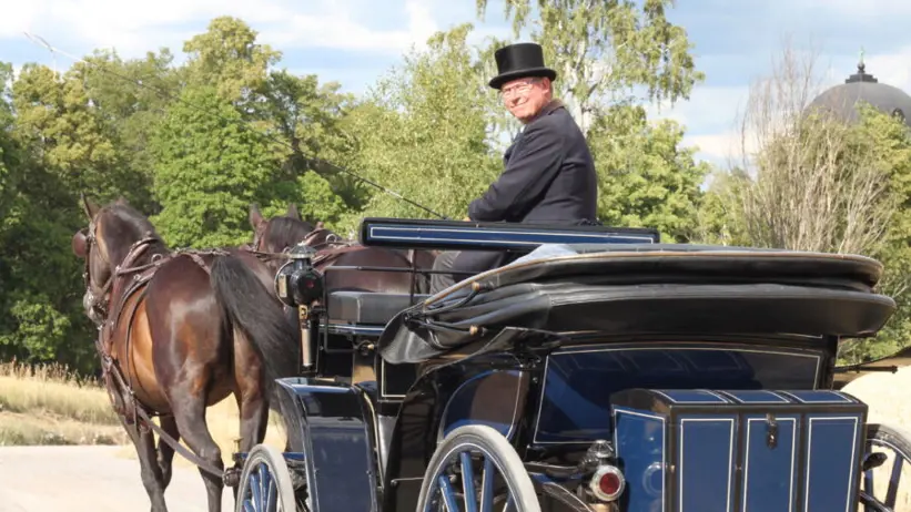 Anders Eriksson som finklädd och med hatt kör en mörkblå hästvagn med två mörkbruna hästar framför.