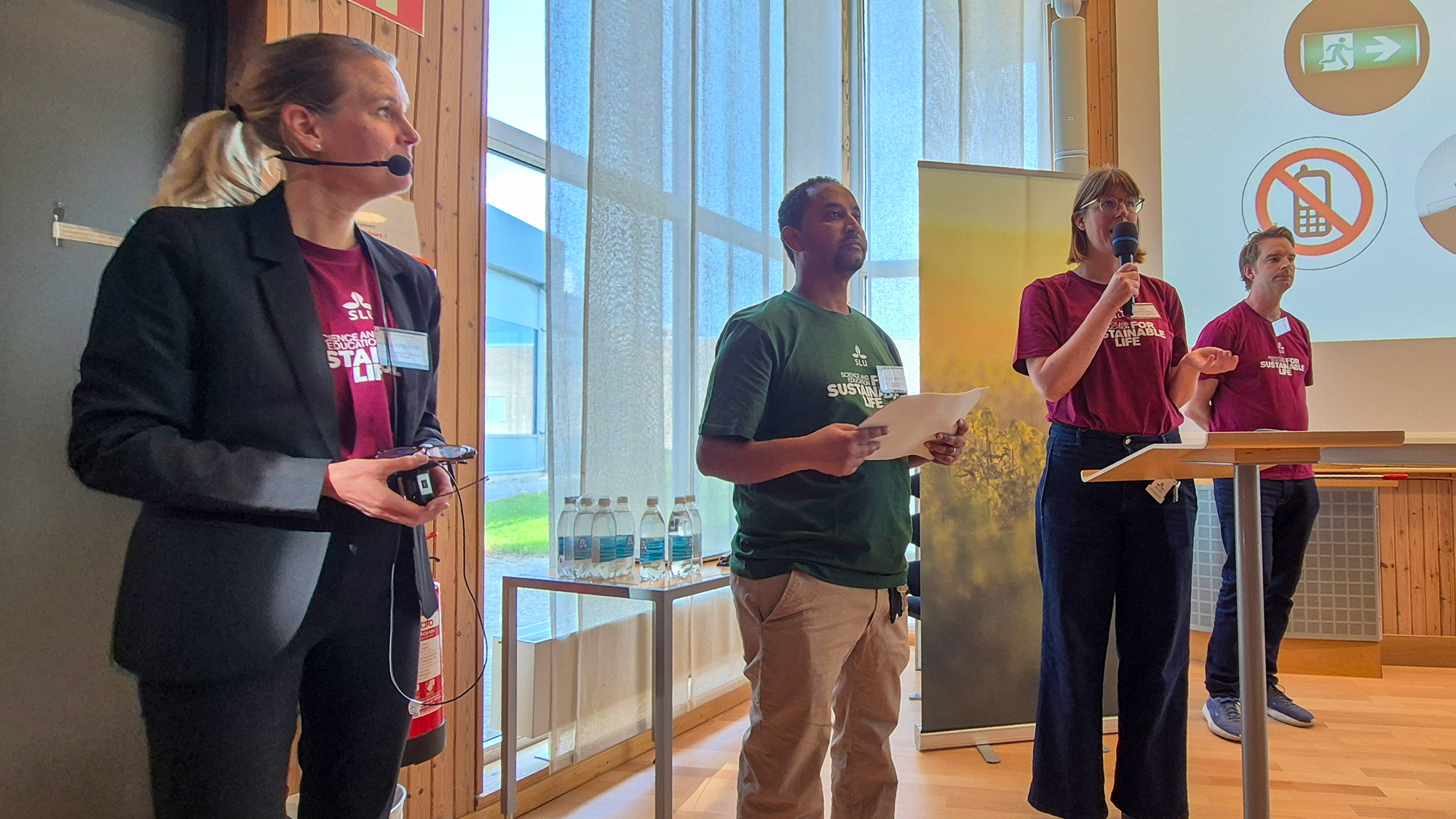 Four people stand in front of an audience with microphones. Photo.