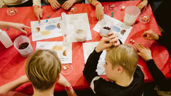 Children sit at a red table with plastic jars of seeds and paper with information. Photo.