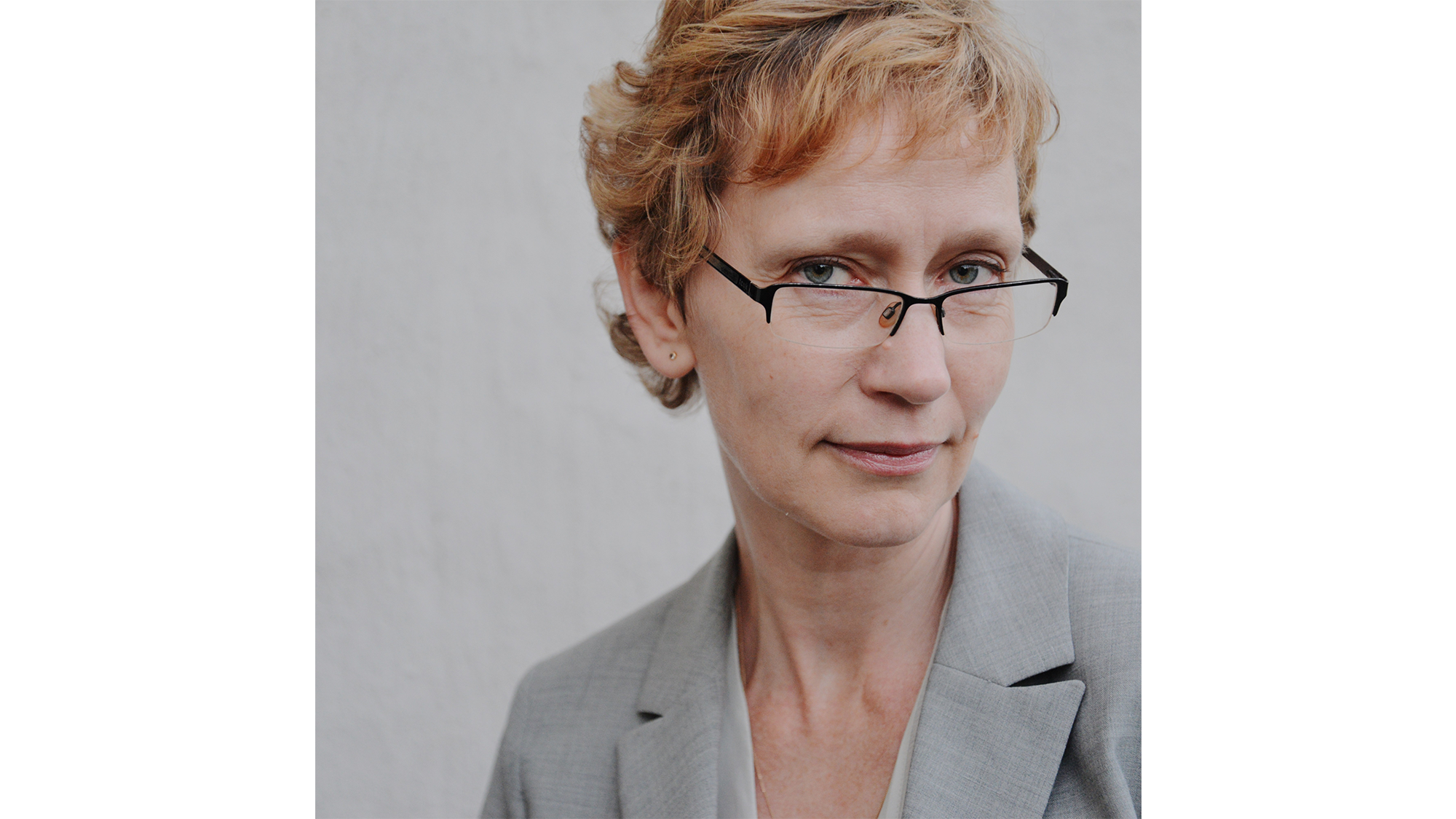 Rödblond kvinna med kort, lockigt hår, glasögon och grå kavaj står mot grå bakgrund. Foto