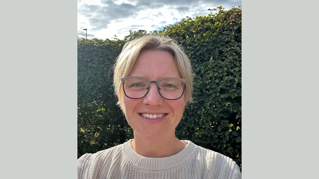 Porträttbild på Maria Gustavsson utomhus med grönt bladverk och blågrå himmel i bakgrunden.