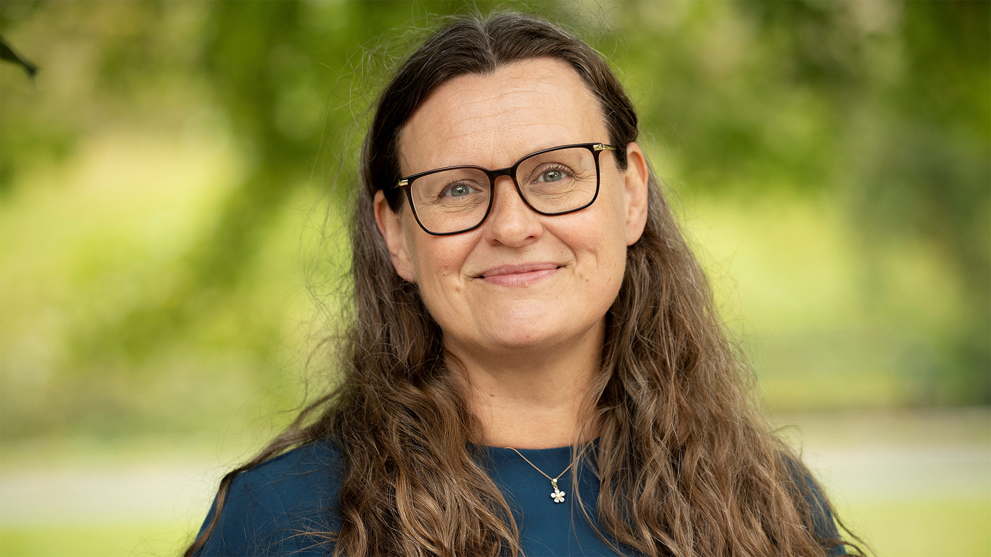 Kvinna med brunt, långt, lockigt hår och glasögon står utomhus med blå tröja. Foto