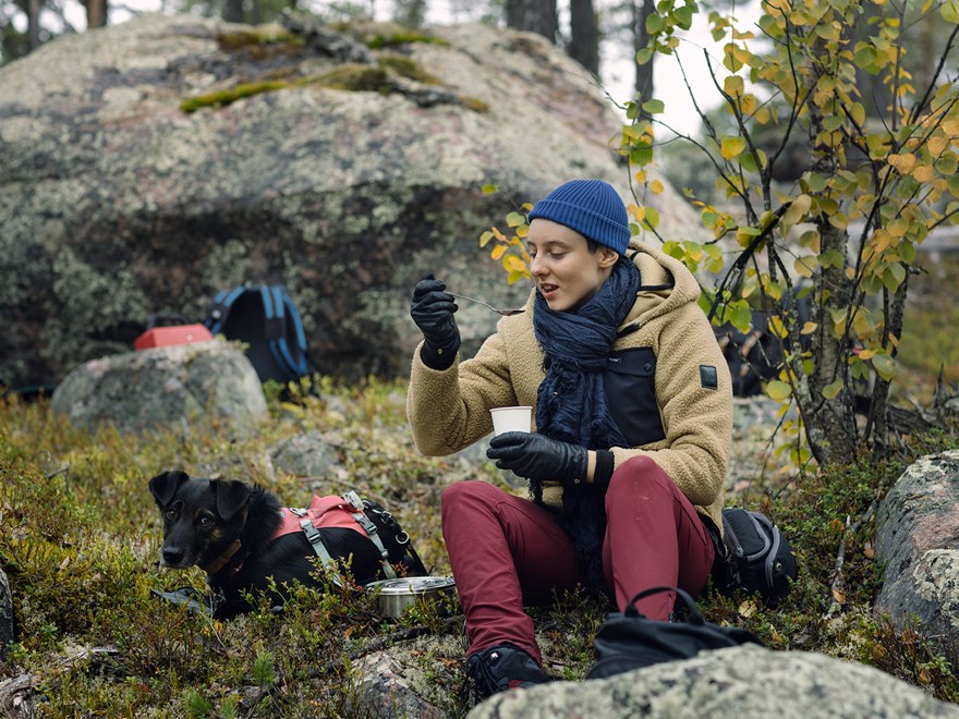 Student som sitter i naturen och äter mat ur en burk. Hund ligger bredvid. Foto.