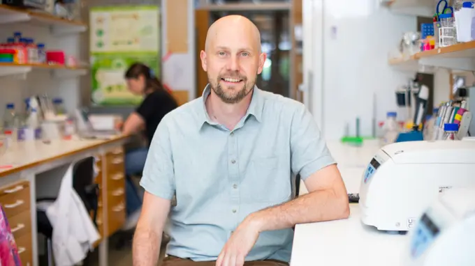 A person with a beard and a short sleaved shirt sitting in a laboratory looking into the camera.
