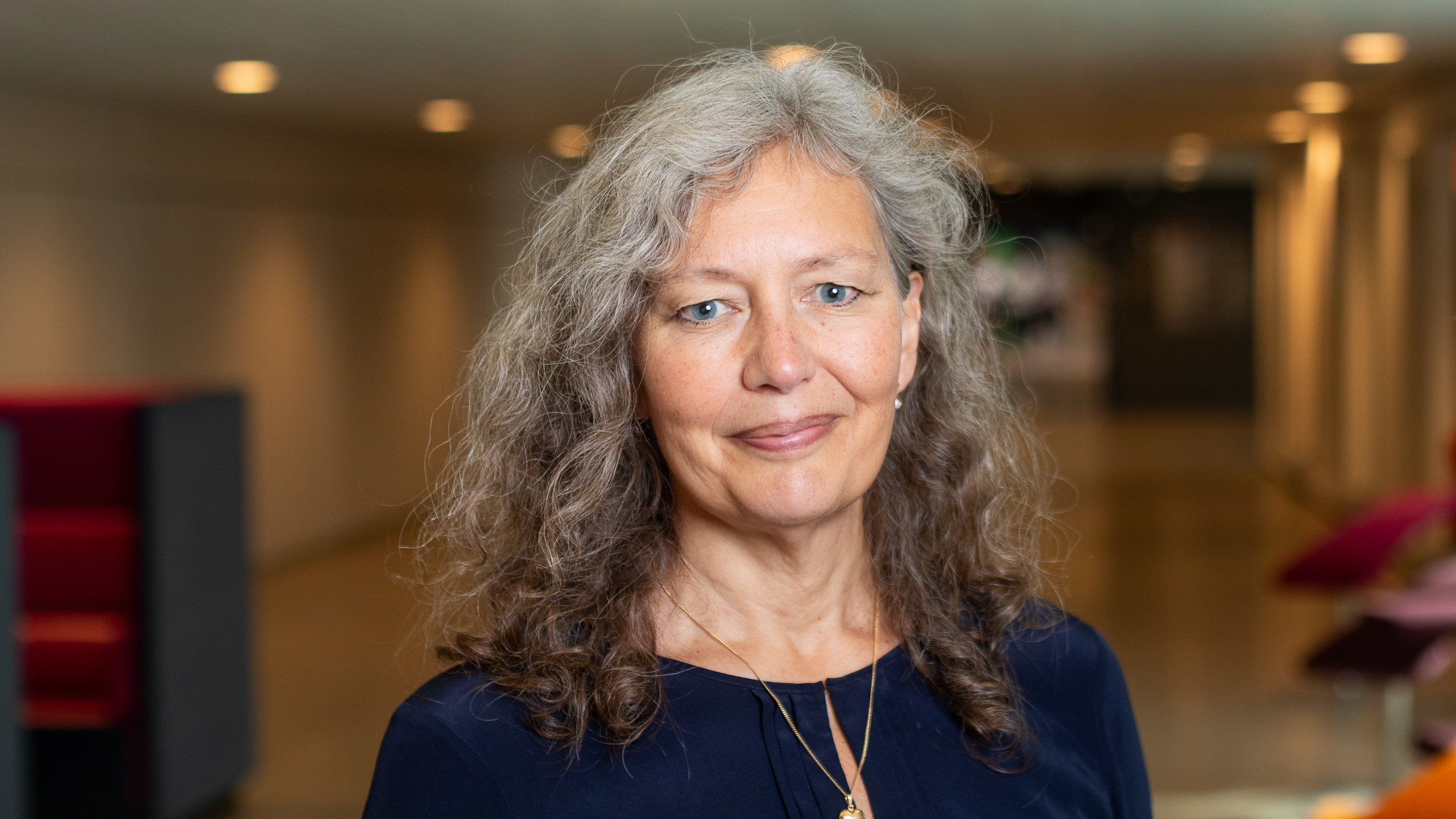 A person with grey, wavy hair stands in a modern indoor setting with soft lighting. The person is wearing a dark blue top and a necklace. In the background, a corridor with red chairs and artwork on the walls is visible.