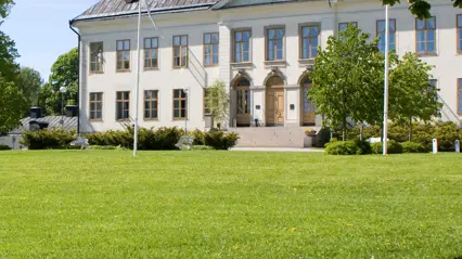 Vitputsad byggnad med trappa och stora fönster omgiven av grönskande parkmiljö, träd och flaggstänger en solig försommardag.